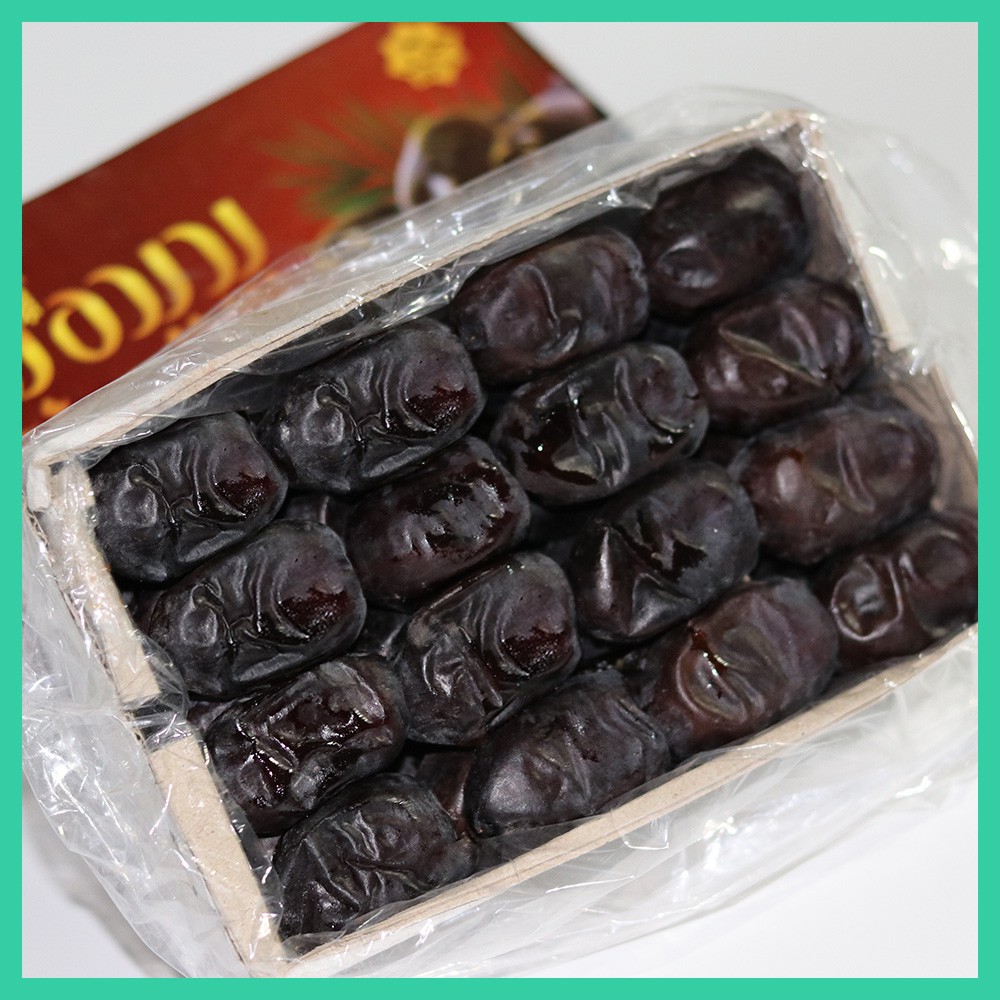 Dulang kurma gelap berkilat disusun rapi dalam kotak kayu nipis dan dibalut plastik ketat, dengan kotak bercetak merah di belakangnya